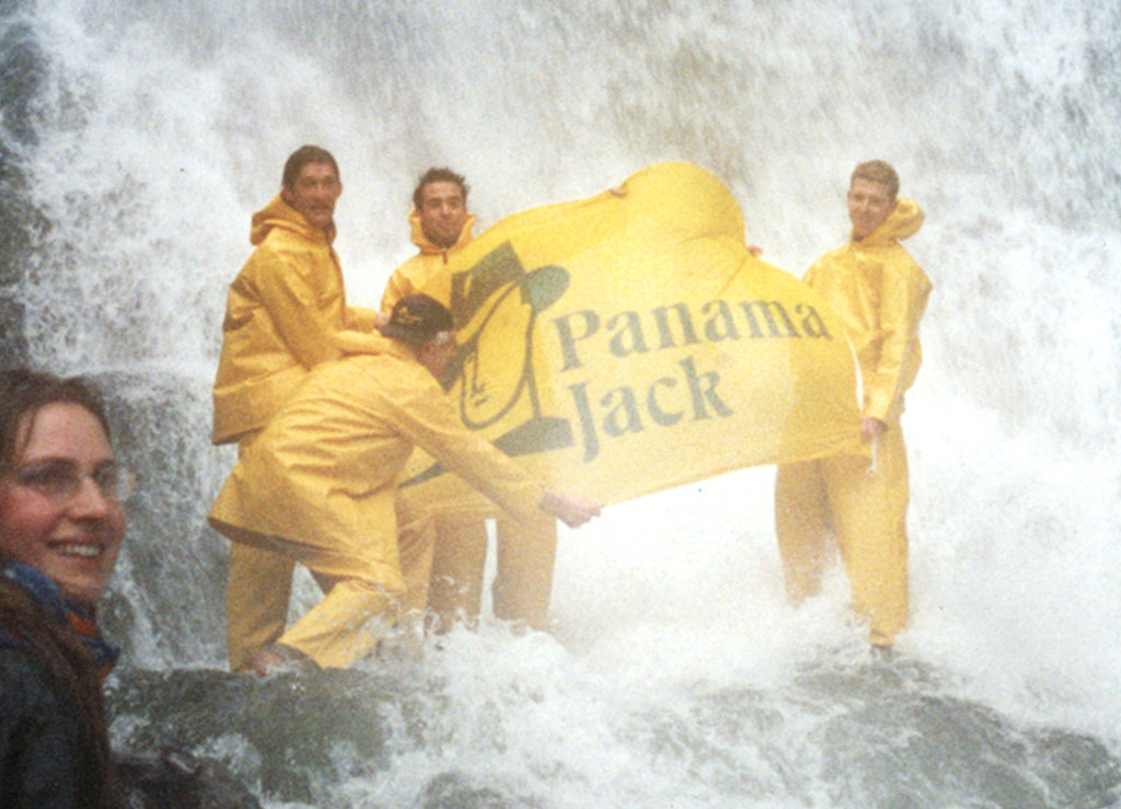 Grupo de jovenes bajo una cascada sujetando una bandera de Panama Jack